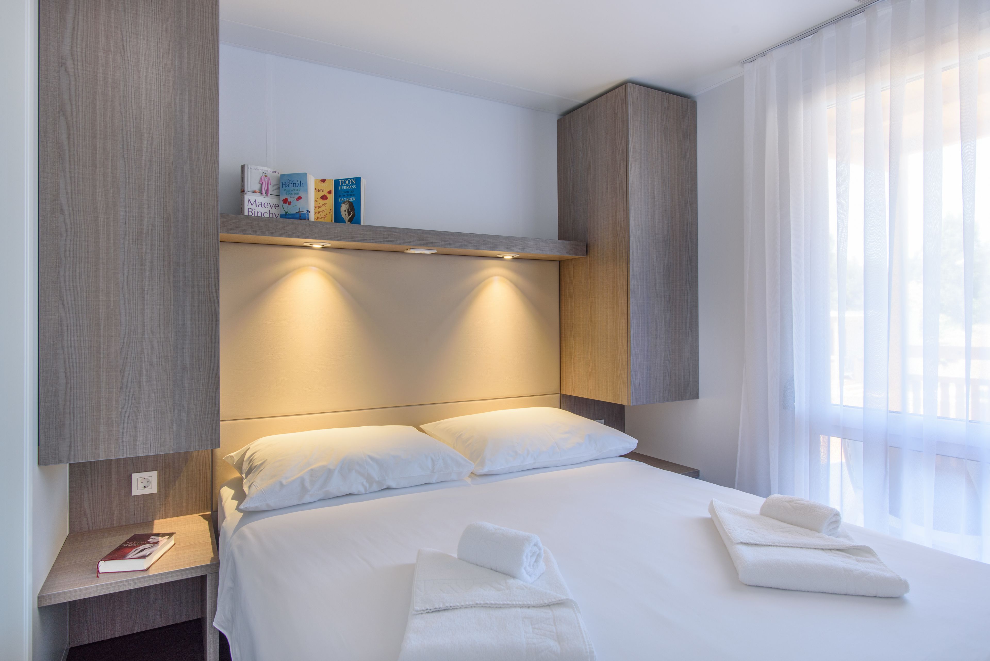 Camera d'albergo moderna con un letto rifatto, un comodino con un libro e una mensola con libri e una rivista.