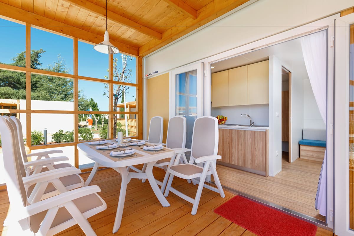 Une cuisine moderne avec des meubles blancs et un évier, un coin repas avec des chaises et une table blanches, et de grandes fenêtres offrant une vue sur l’extérieur.