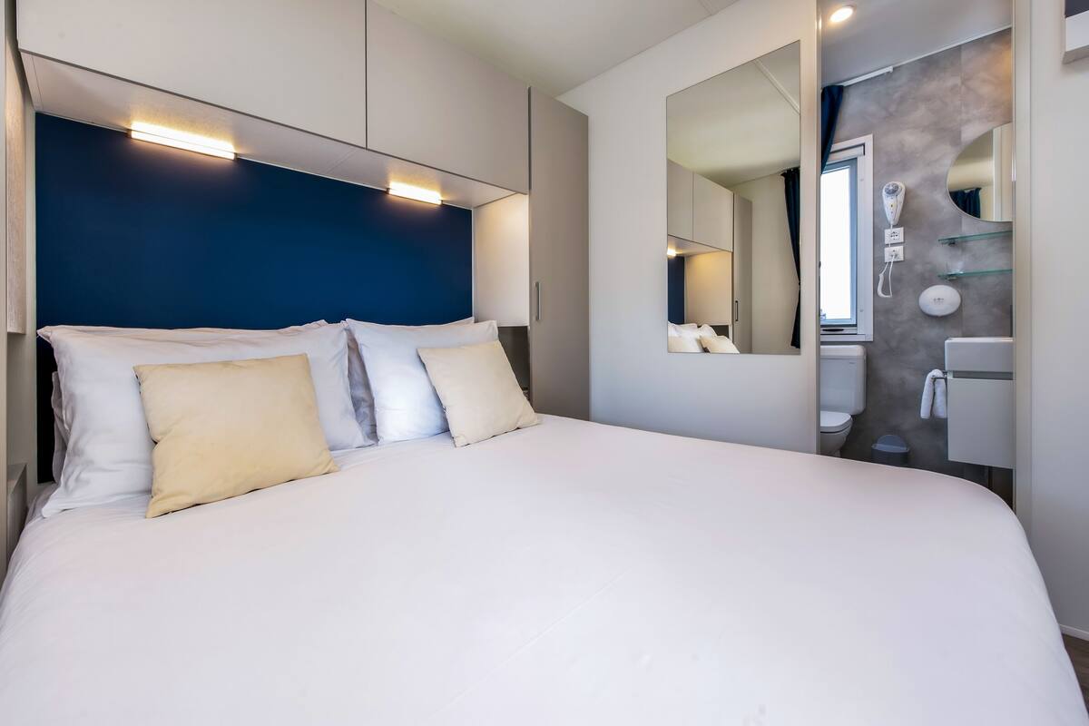 Une chambre d’hôtel moderne dotée d’un grand lit, d’une tête de lit avec éclairage intégré, d’un miroir et d’une salle de bain comprenant des toilettes et un lavabo.