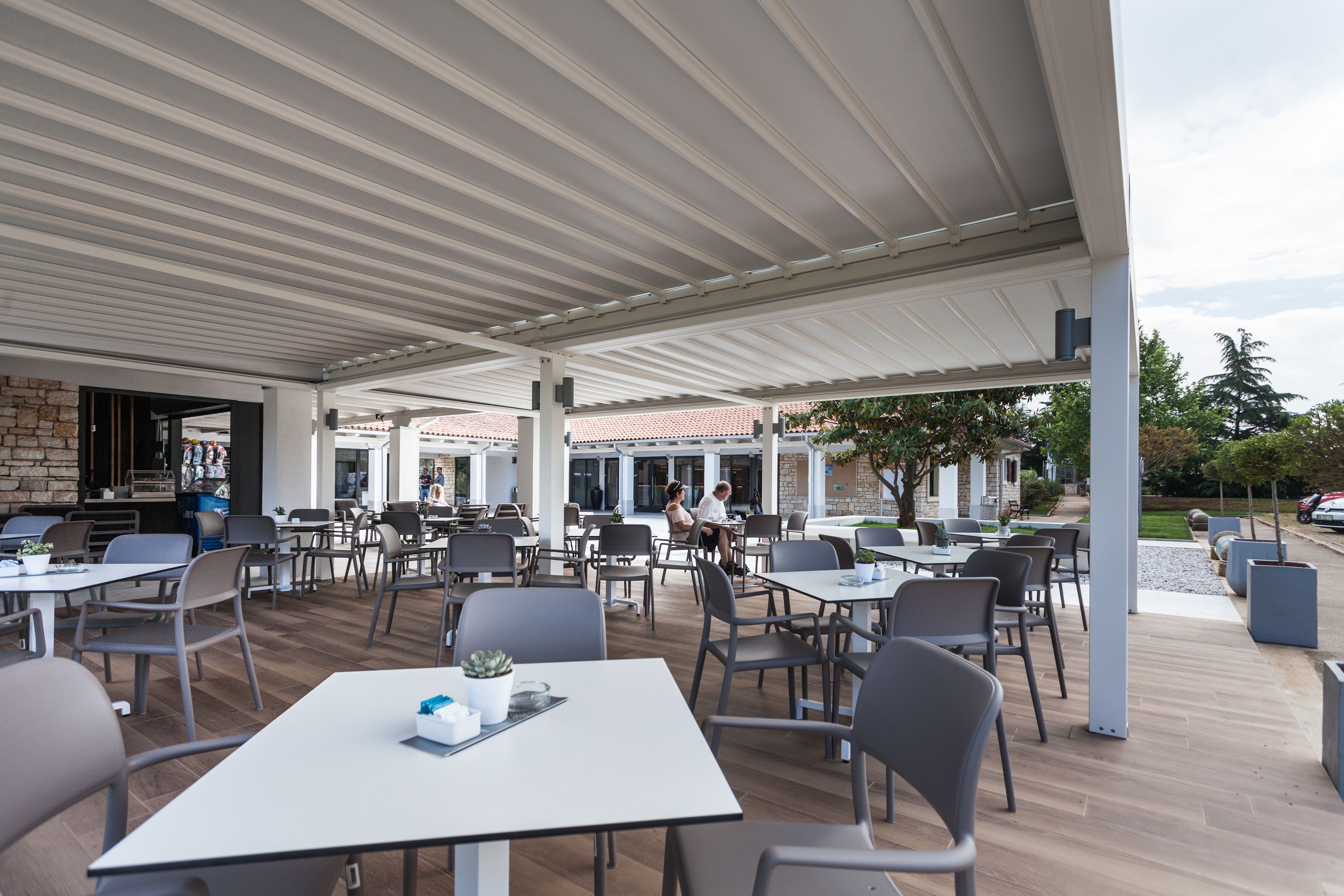 Un cafè a l'aire lliure amb taules i cadires, una paret de pedra i una zona coberta amb teulada de rajola.