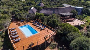 An aerial view of a resort with a wooden deck, a swimming pool, lounge chairs, and thatched-roof buildings. - Woodbury Tented Camp - Amakhala Game Reserve (Sidbury)
