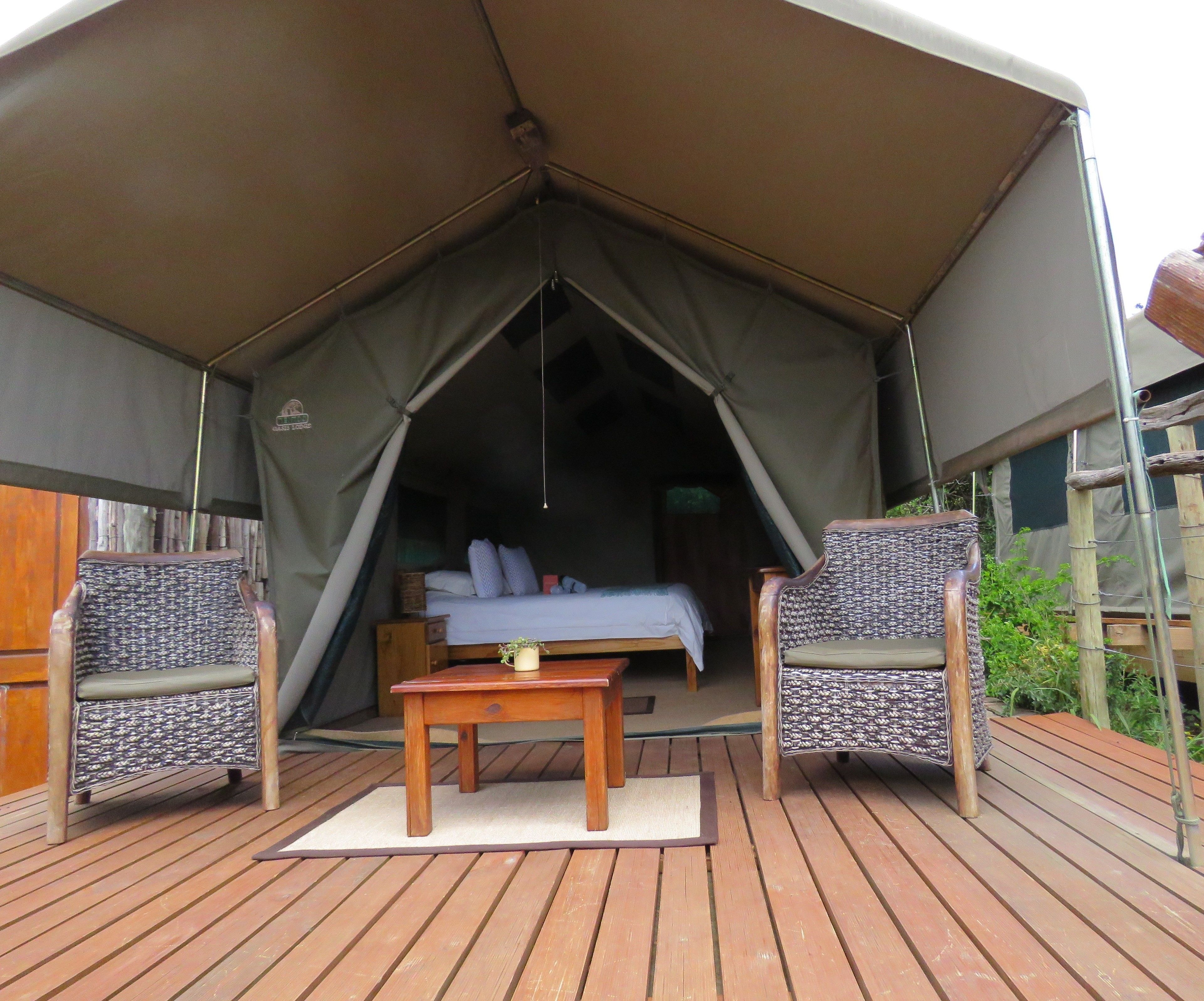 A tented accommodation with a bed, two wicker chairs, a small table, and a wooden deck.