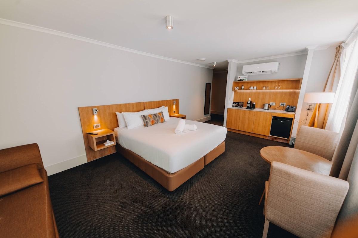 A hotel room with a bed, bedside table, chair, and kitchenette.