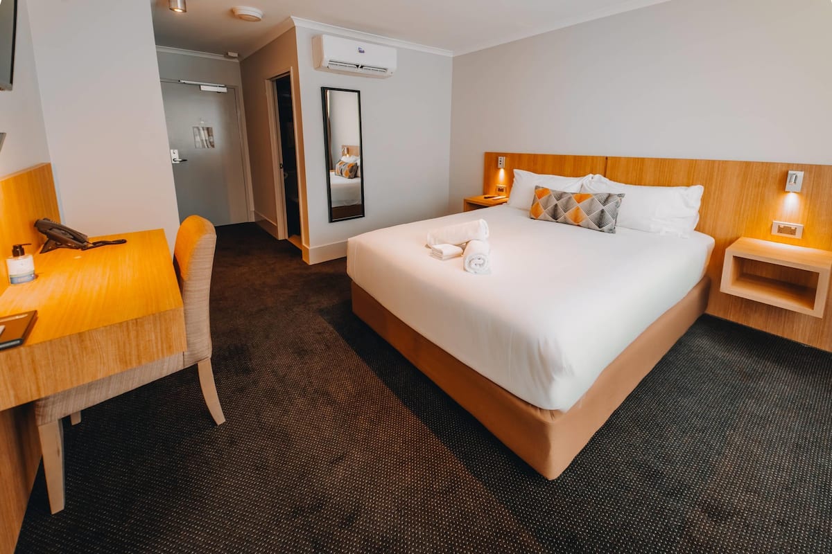 A hotel room with a bed, a desk, a chair, a mirror, a bedside table, and an air conditioning unit.