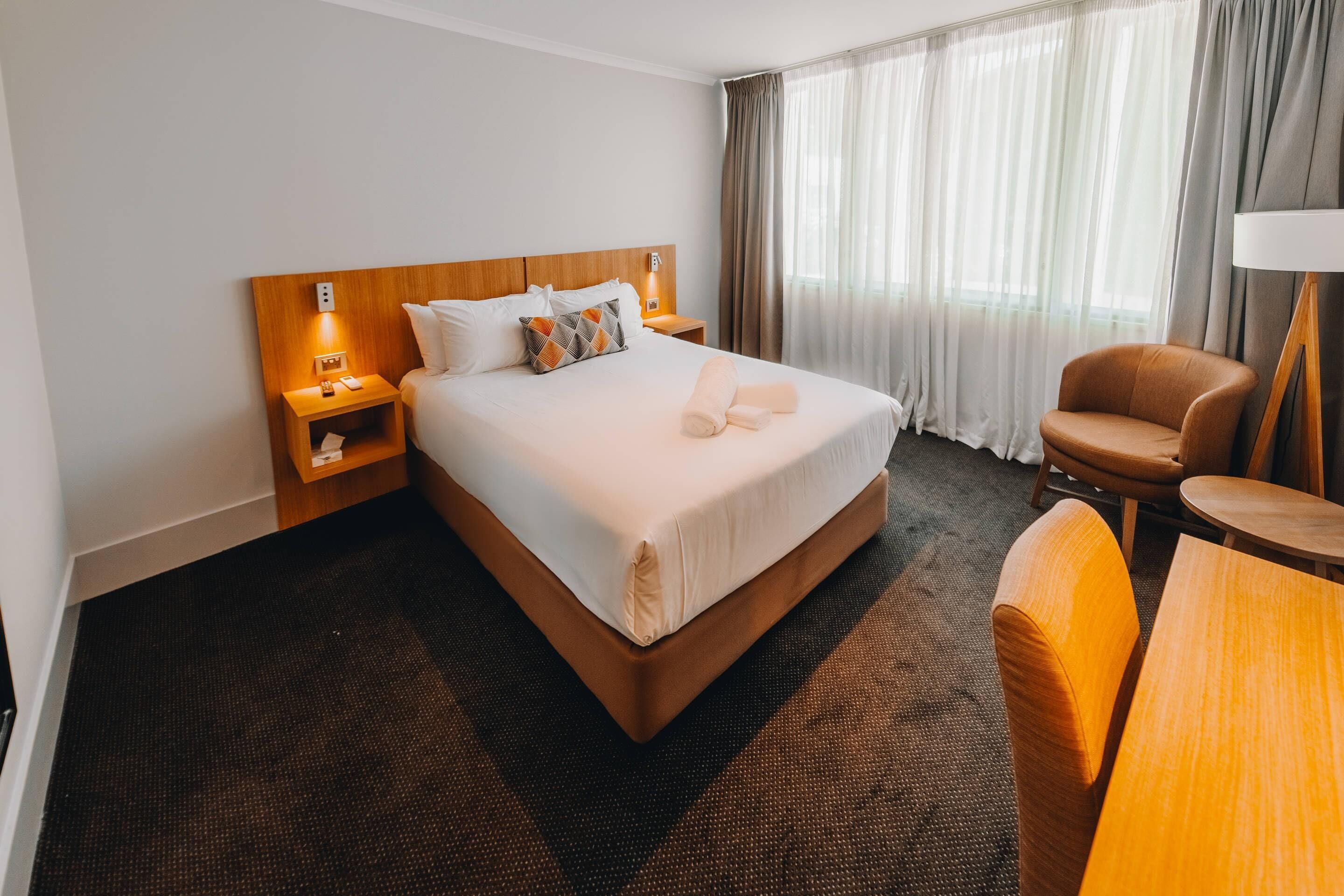 A hotel room with a large bed, a nightstand, a chair, a table, and a lamp.