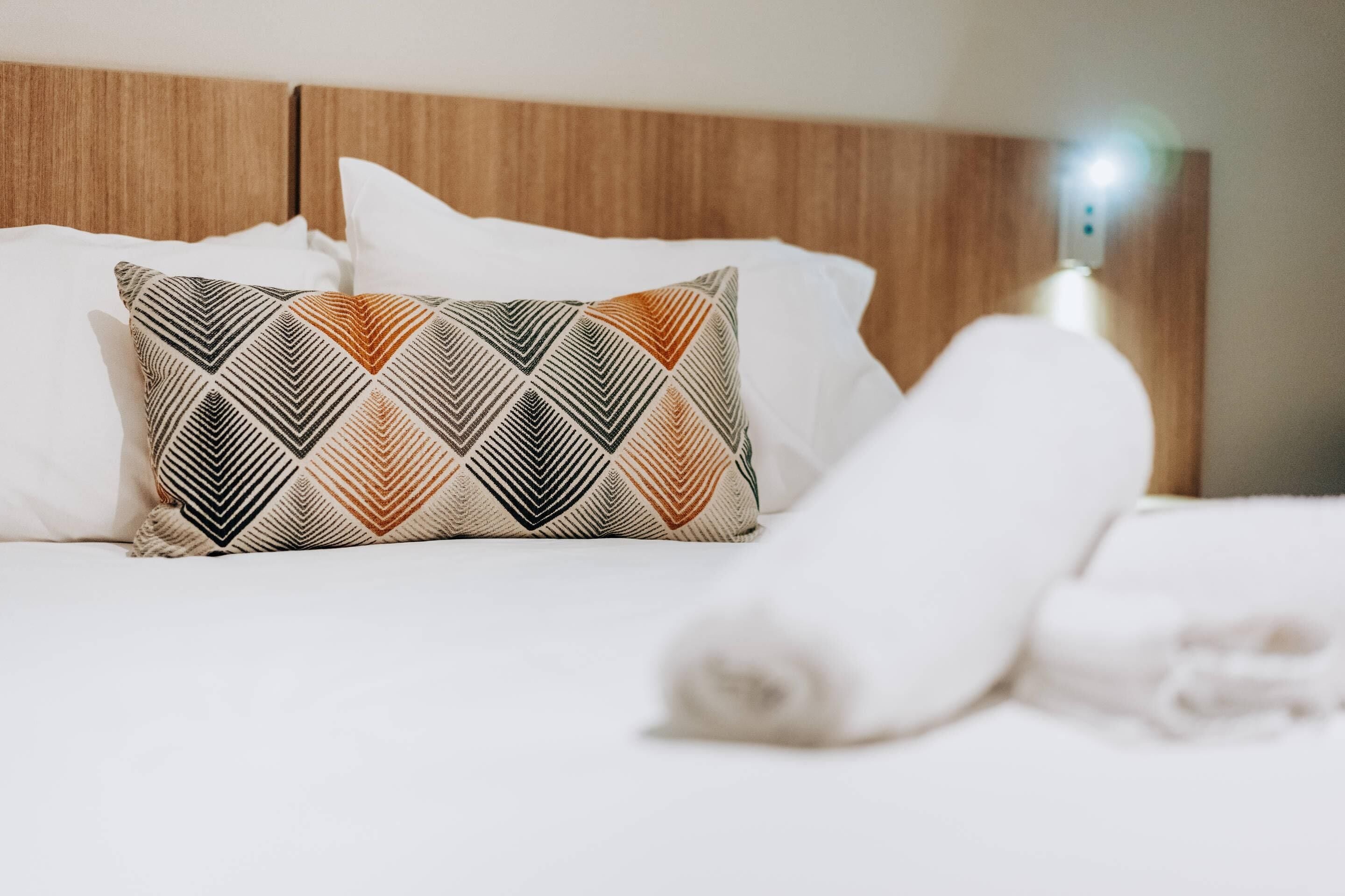 A neatly made bed with a patterned pillow and a rolled white towel.