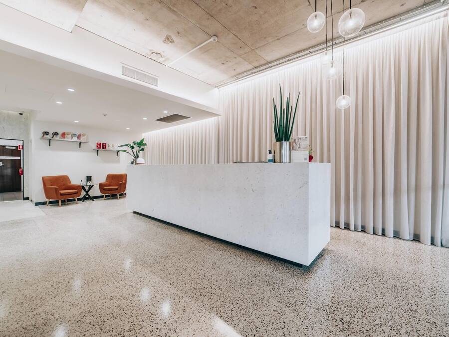 A modern reception area with a white reception desk, two orange chairs, a small table, and a potted plant.