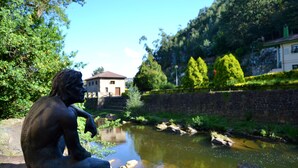 Une statue représentant une figure assise, placée près d’une rivière, avec un mur de pierre et des arbres en arrière-plan.