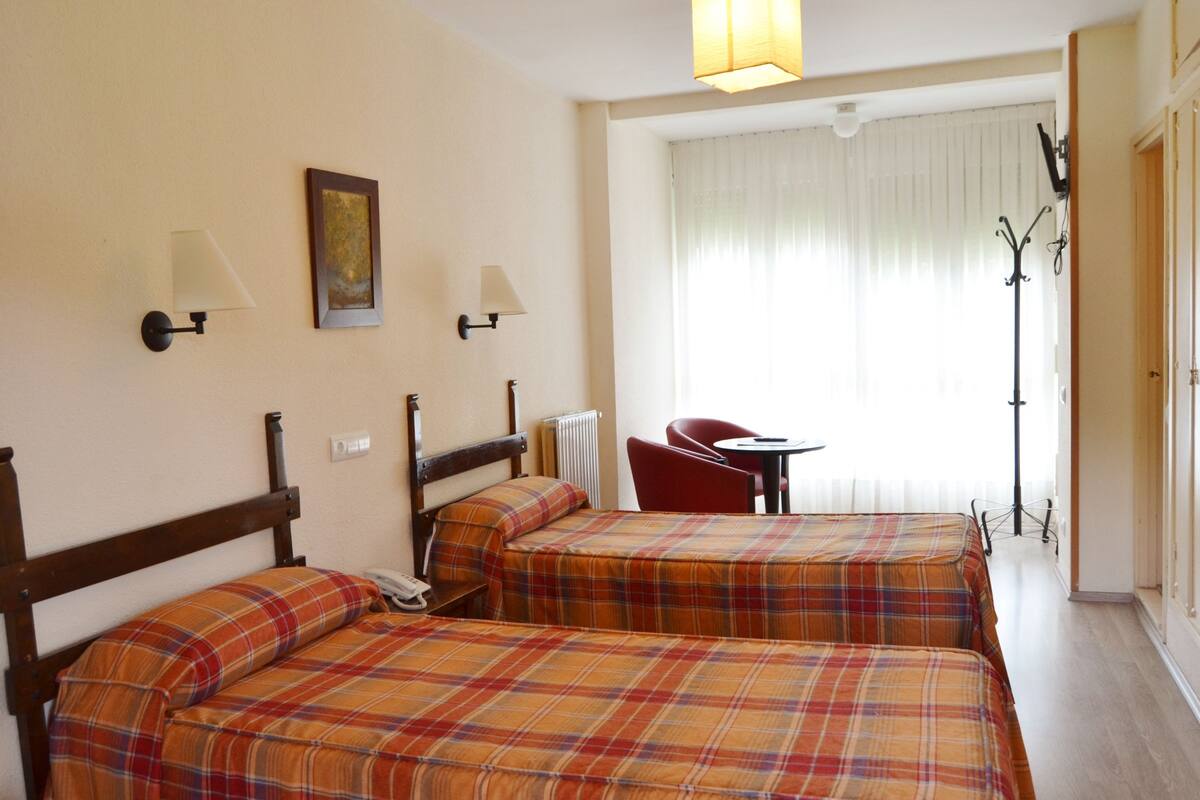Ein Hotelzimmer mit zwei Betten, einem kleinen Tisch, einem Sessel und Blick ins Freie durch ein Fenster.