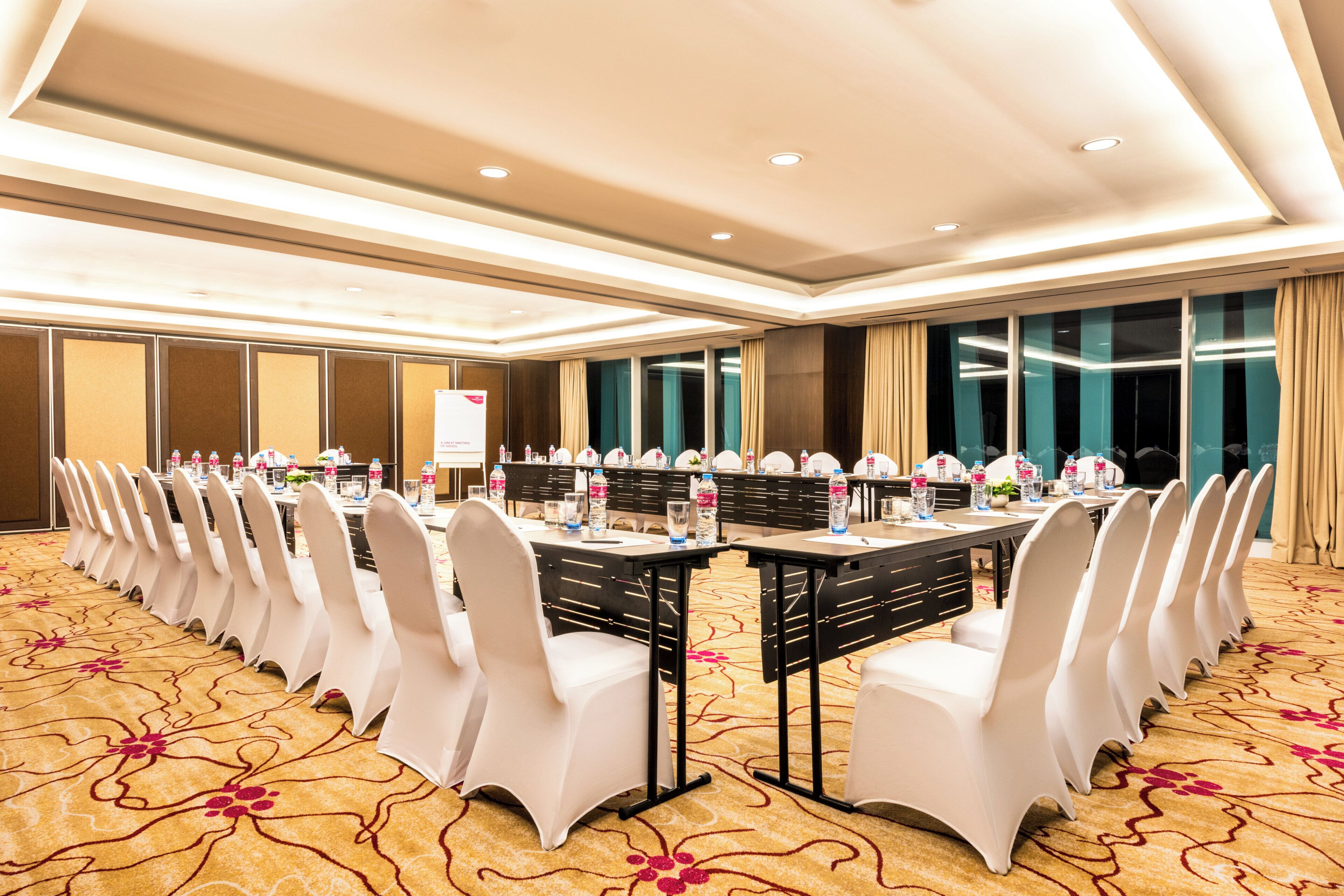 A conference room with long tables, white chairs, and a carpeted floor.