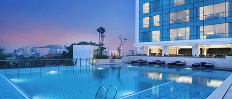 A modern hotel with a rooftop infinity pool, lounge chairs, and a view of the city skyline.