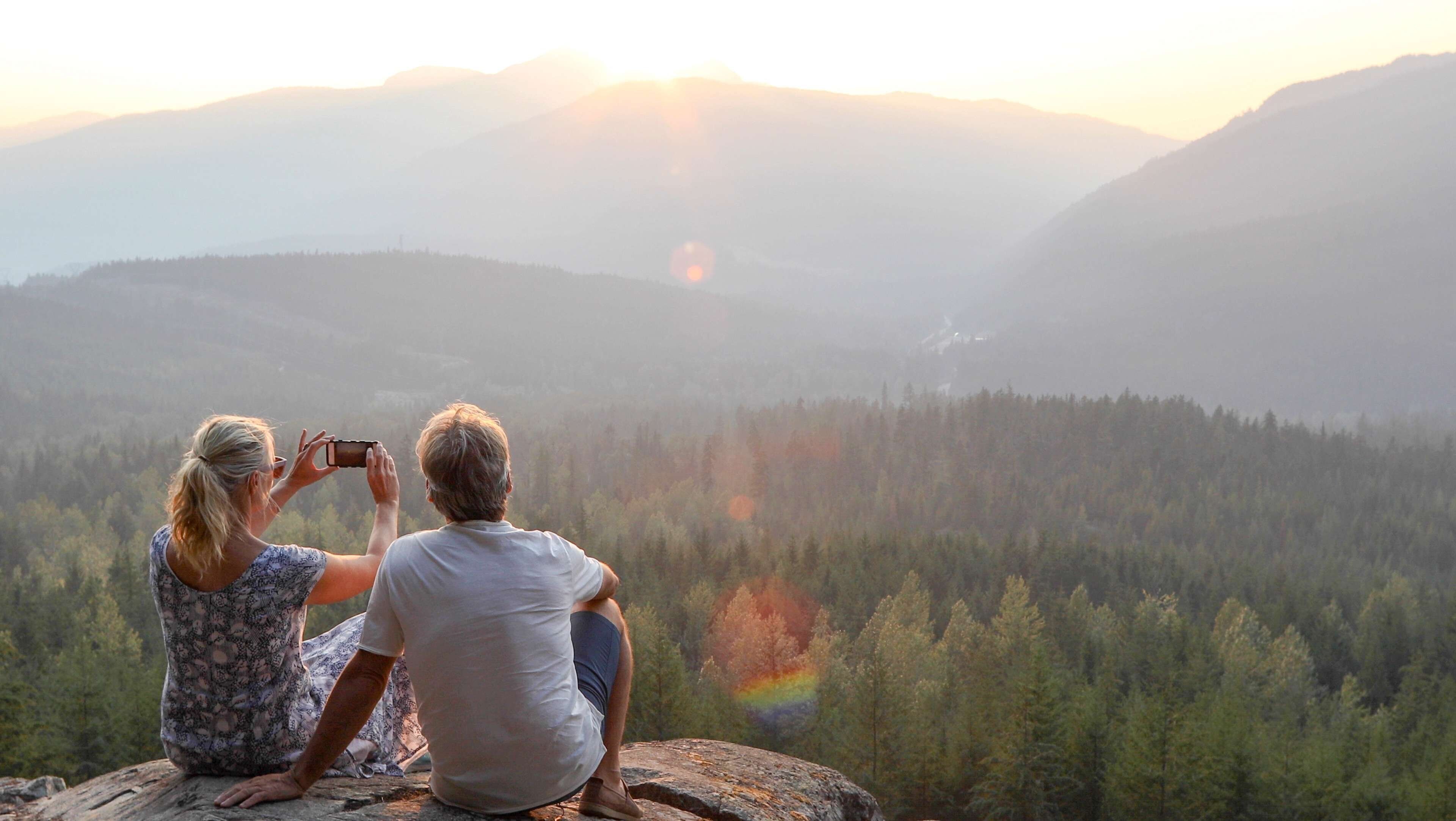 Two people sitting on a rock with a forest and mountains in the background.