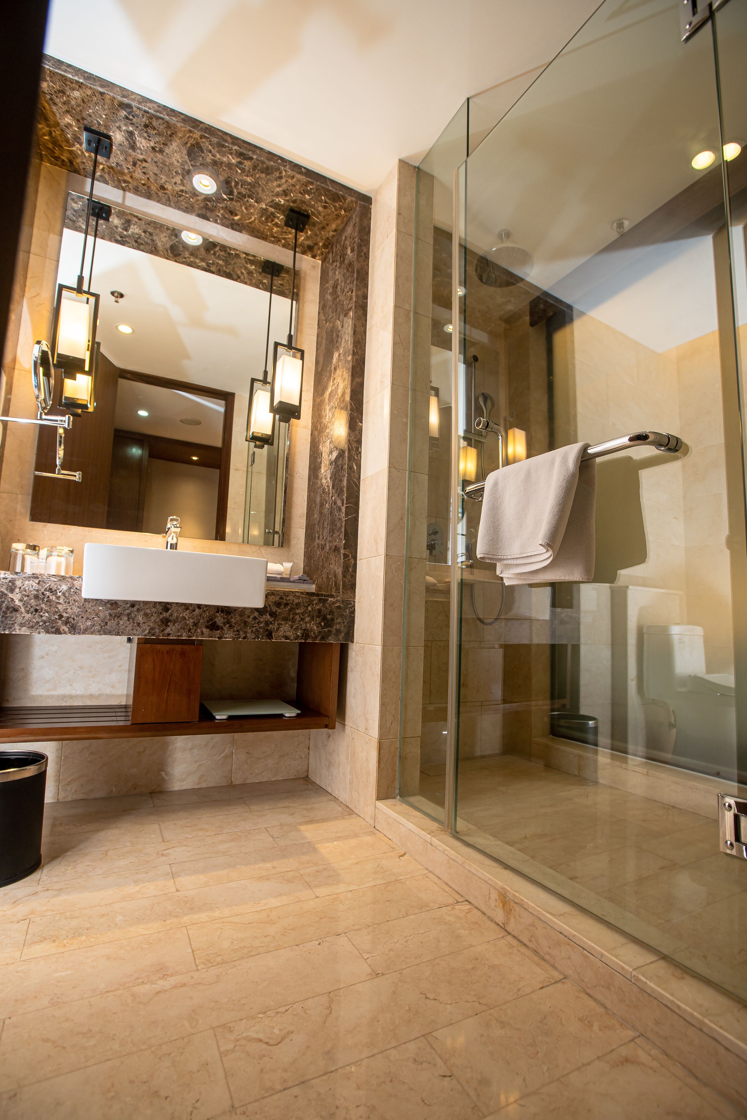 A modern bathroom with a glass-enclosed shower, a marble-tiled floor, and a large mirror with pendant lighting.