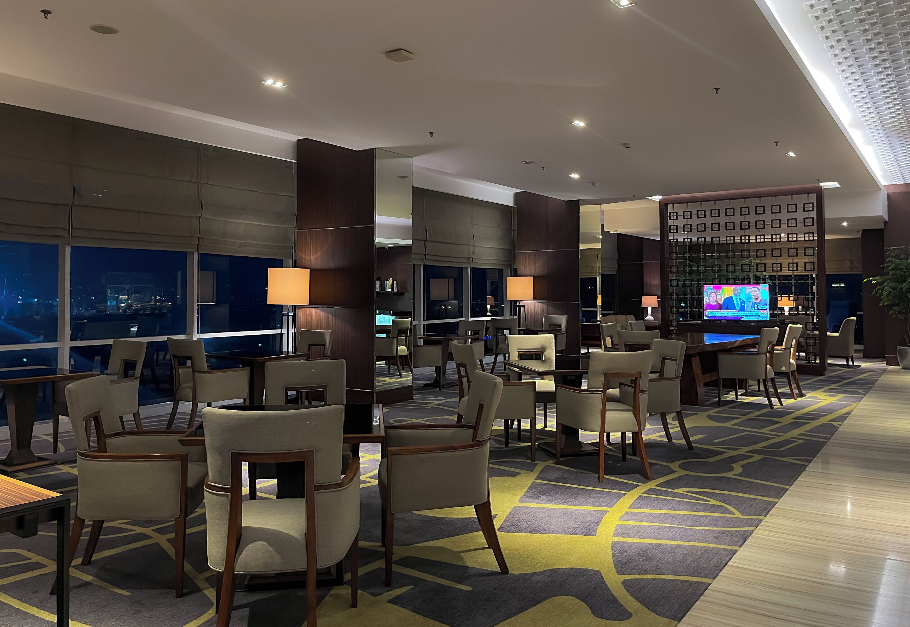 A hotel lobby with multiple seating areas, a large television screen, and a view of the city at night.