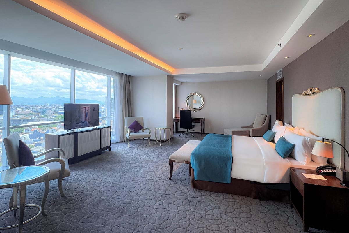 A hotel room with a large bed, a desk, a chair, a TV, and a view of the city.