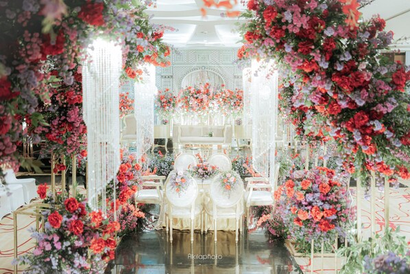 Un espacio interior con un pasillo central, sillas blancas y decoración floral.