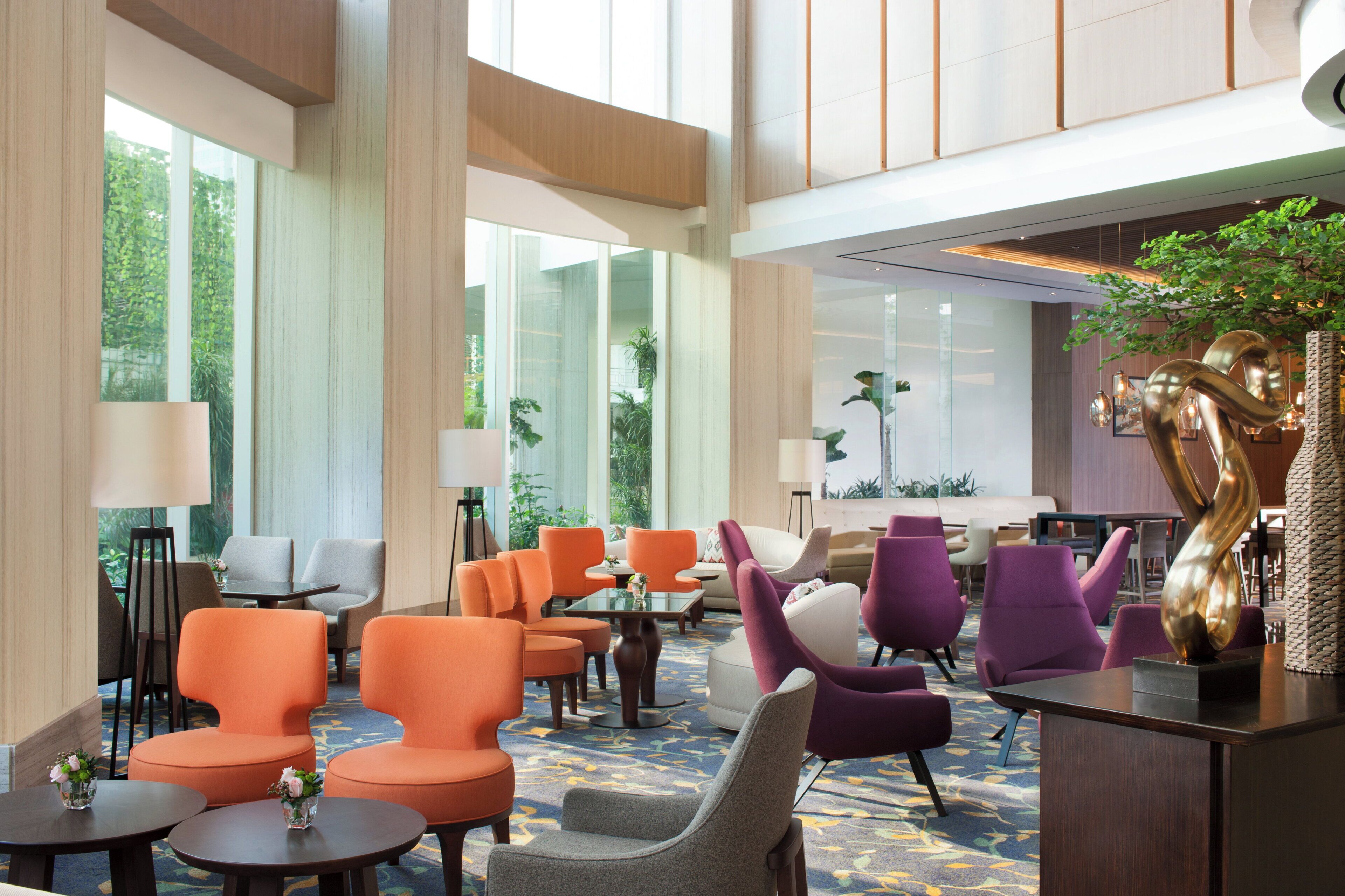 Eine großzügige Lobby mit hohen Decken, großen Fenstern und verschiedenen Sitzgelegenheiten, darunter orangefarbene und violette Stühle.