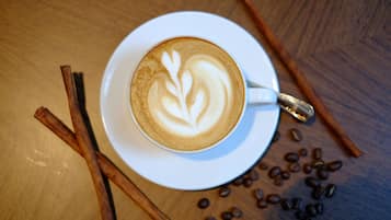 Una taza de café con arte de latte sobre un platillo blanco, rodeada de granos de café y palitos de canela sobre una mesa de madera.