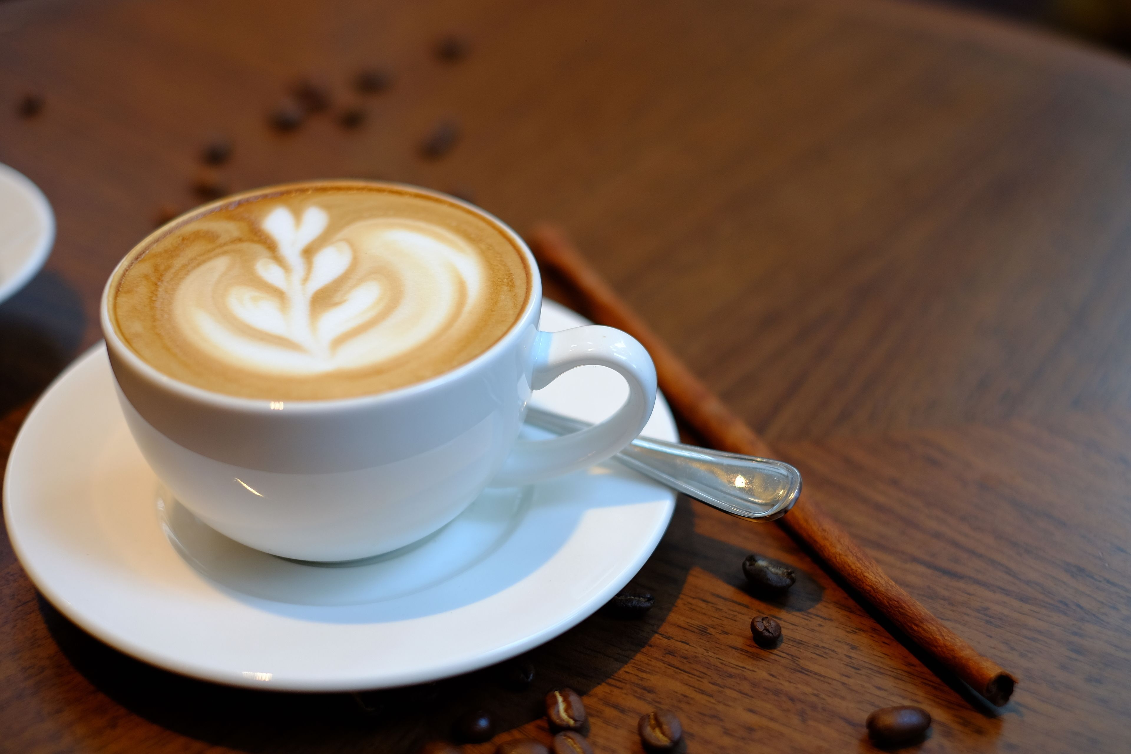 Une tasse de café blanc, décorée d’un motif en latte art, posée sur une soucoupe, le tout placé sur une table en bois parsemée de grains de café et agrémenté d’une tige de cannelle.