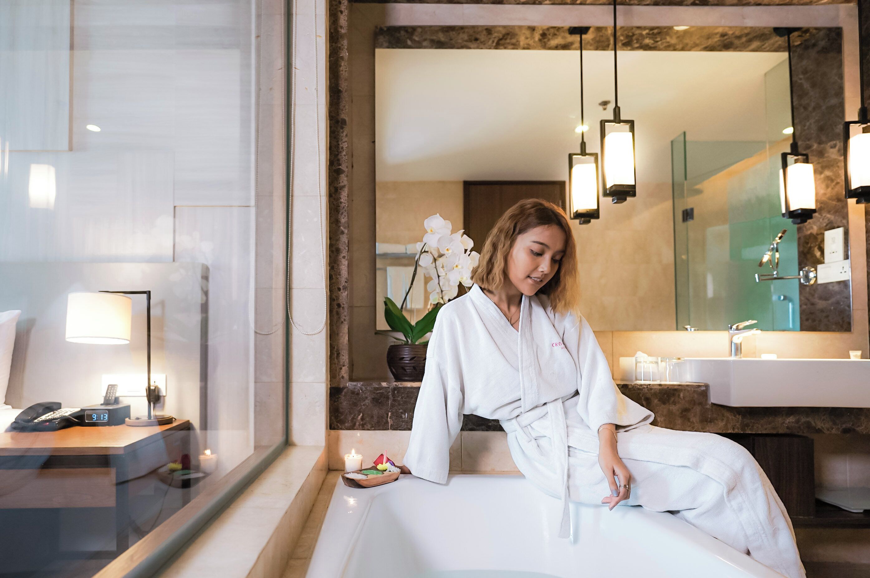 Une salle de bain d’hôtel avec une baignoire, un miroir et un lavabo. Il y a une horloge au mur et une plante sur le comptoir.