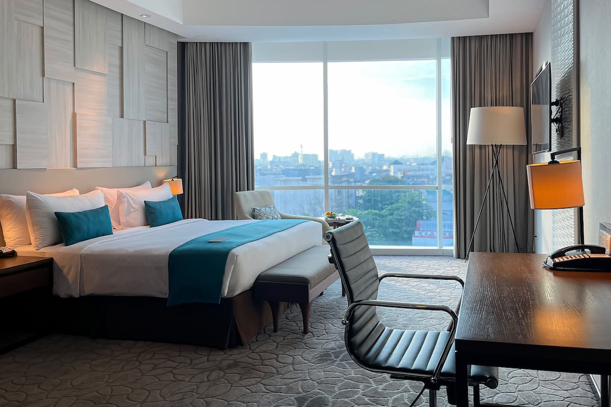 A hotel room with a large bed, a desk, a chair, a nightstand, and a view of the city through large windows.