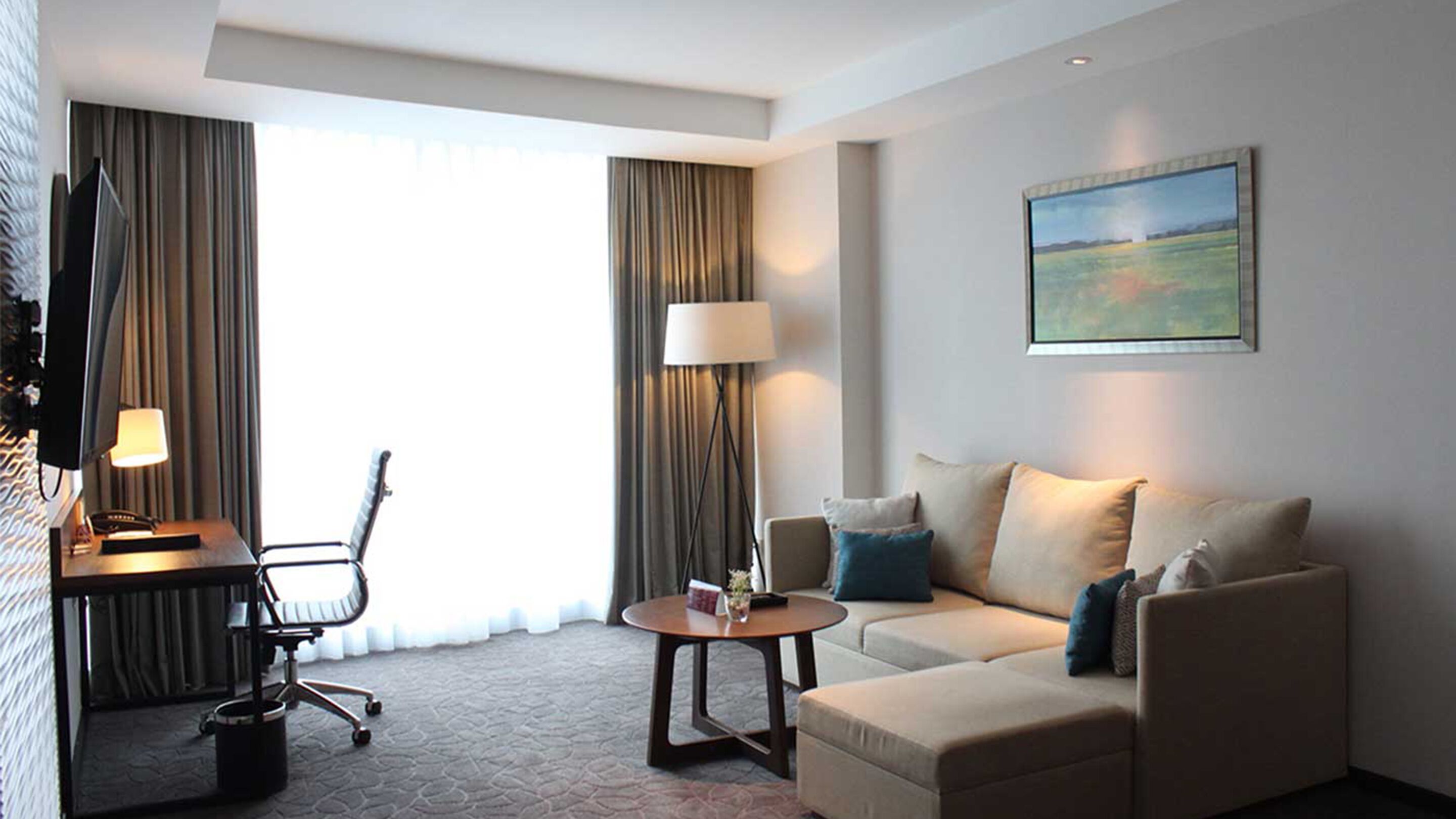 A hotel room with a sofa, a desk, a chair, a lamp, and a painting on the wall.
