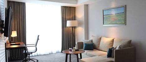 A hotel room with a sofa, a desk, a chair, a lamp, and a painting on the wall.