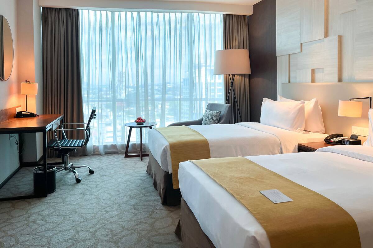 A hotel room with two beds, a desk, a chair, a small table with a bowl of fruit, and a view of the city through large windows.