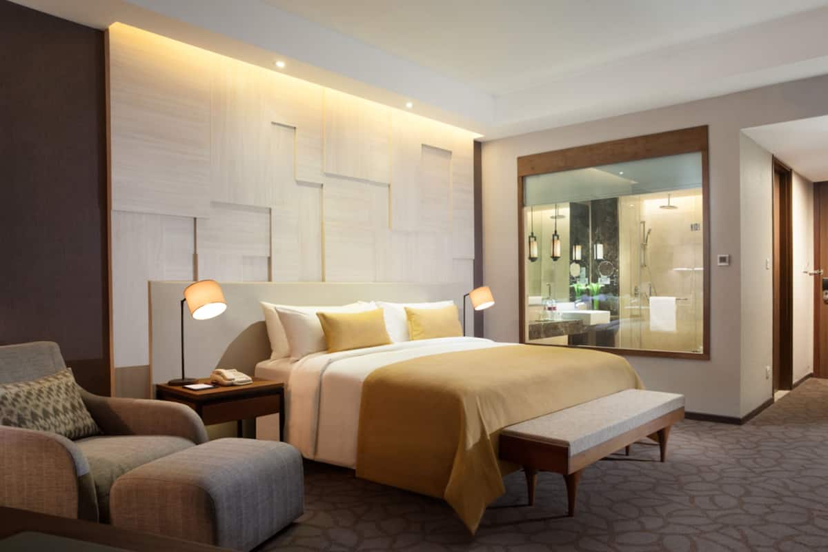 A modern hotel room with a large bed, bedside lamps, a bench, a chair, and a large mirror reflecting the bathroom.