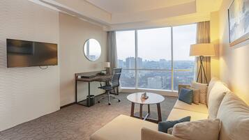 A hotel room with a large window, a flat-screen TV mounted on the wall, a desk with a chair, a sofa, and a small round table.