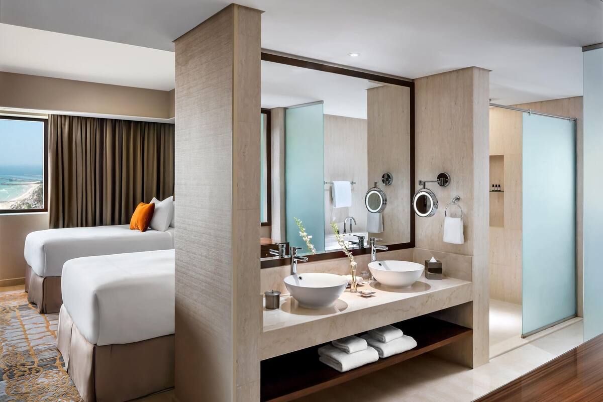 A modern hotel room with two beds, a large bathroom with a vanity, and a view of the ocean.