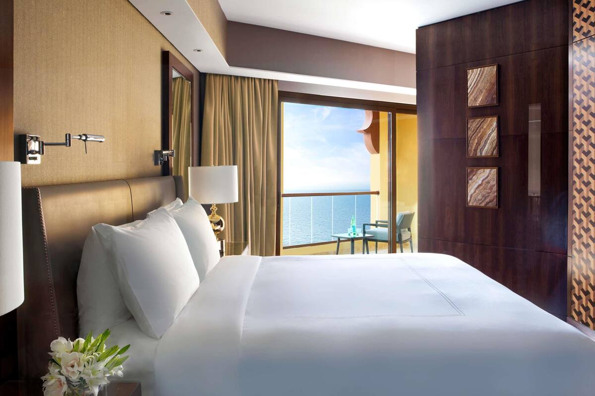 A hotel room with a large bed, a nightstand with a lamp, a vase of flowers, a view of the sea, and a balcony with a table and chair.