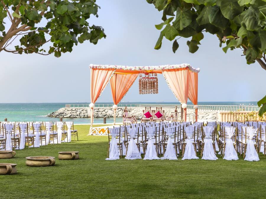 Een bruiloft buiten met een podium, stoelen en tafels op het gras bij een strand.