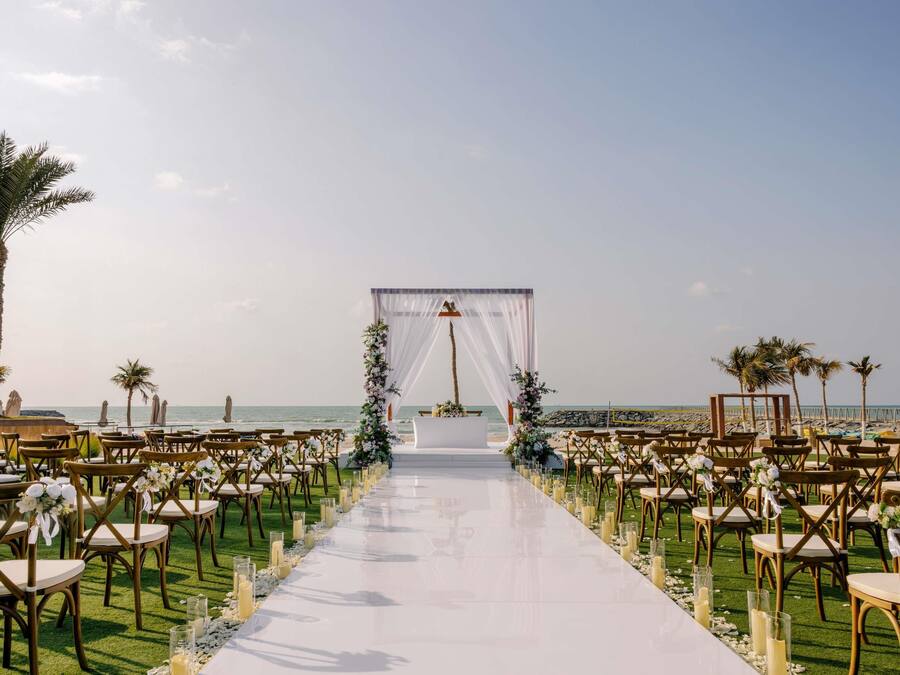 Allestimento per un matrimonio sulla spiaggia, con una navata bianca, decorazioni floreali e sedie in legno.