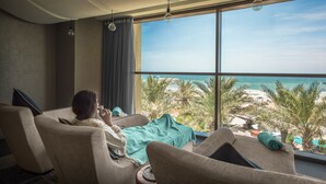 A room with a large window overlooking a beach, a person sitting on a chair, and a view of palm trees and the ocean. - Fairmont Ajman (Ajman)