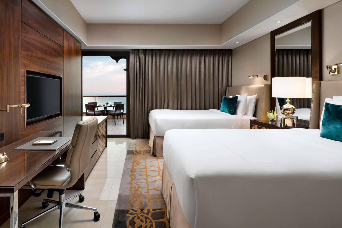 A hotel room with two beds, a desk with a chair, a television, and a view of the ocean.