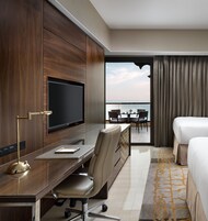Habitación de hotel con una cama grande, un escritorio con televisor, una silla y vista al mar desde un balcón.