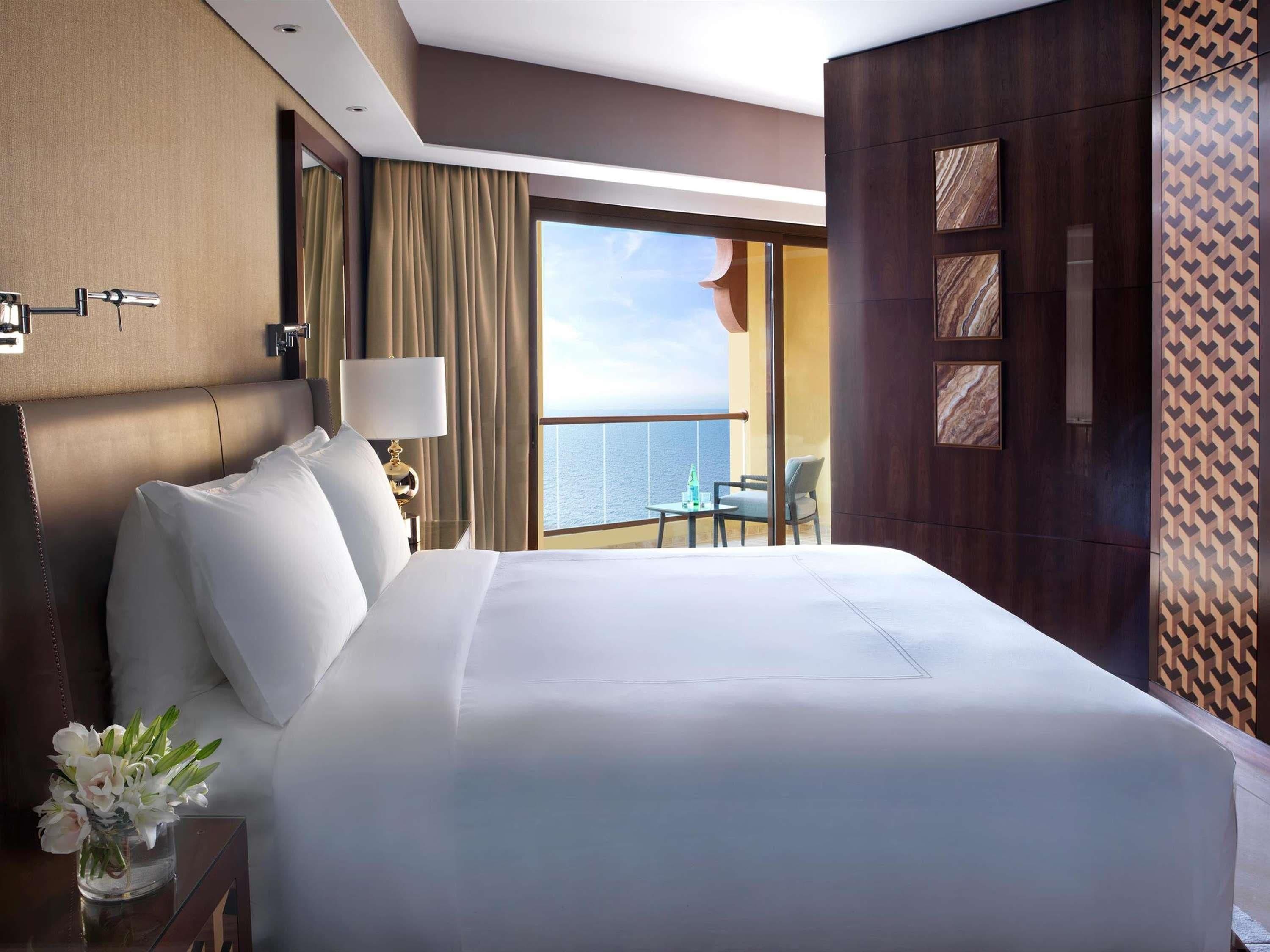 Habitación de hotel con una cama grande, una mesita de noche, una lámpara, un jarrón con flores y vistas al mar, además de un balcón con mesa y sillas.