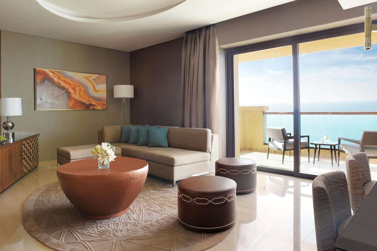 A modern hotel room with a large window offering a view of the sea, a sofa, a coffee table, and a dining area with chairs.