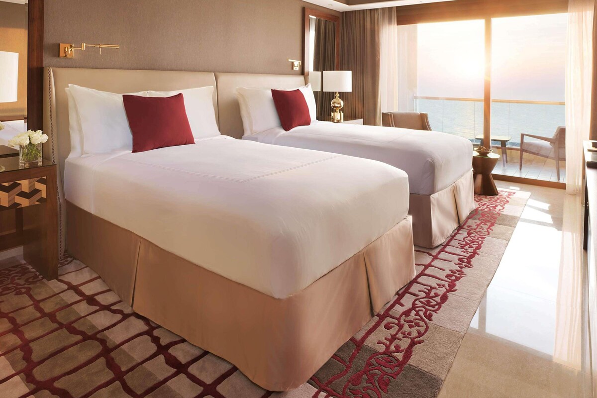 A hotel room with two beds, a large window with a view, a nightstand, and a lamp.