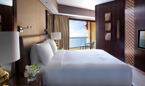 A hotel room with a large bed, a nightstand with a lamp, a vase with flowers, and a view of the sea through a large window. - Fairmont Ajman (Ajman)