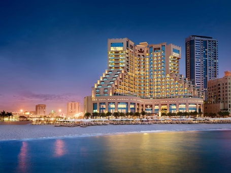 A multi-story hotel with balconies, a pool area with sun loungers, and a beachfront view.