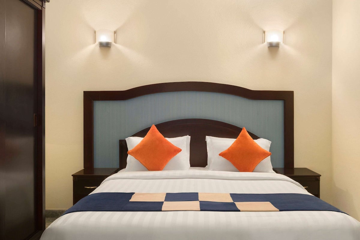 Un lit bien fait, recouvert d’une couverture à carreaux bleus et blancs, et agrémenté de deux coussins orange. Deux tables de chevet avec des lampes se trouvent de part et d’autre du lit, et les murs sont peints dans une couleur beige clair.