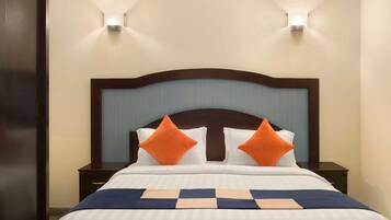 Un lit bien fait, recouvert d’une couverture à carreaux bleus et blancs, et agrémenté de deux coussins orange. Deux tables de chevet avec des lampes se trouvent de part et d’autre du lit, et les murs sont peints dans une couleur beige clair.