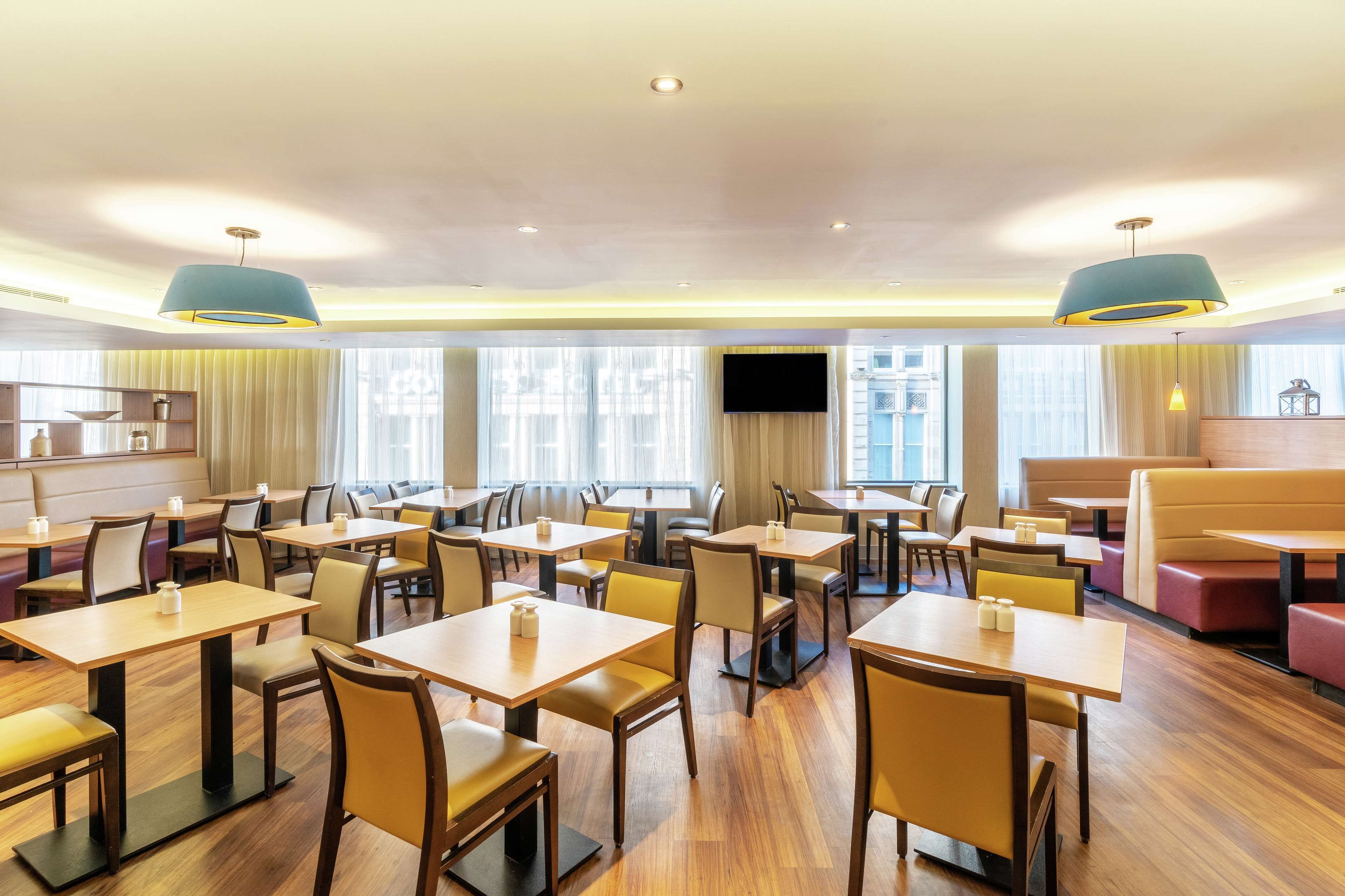 A hotel dining area with multiple tables and chairs, large windows, and ceiling lights.