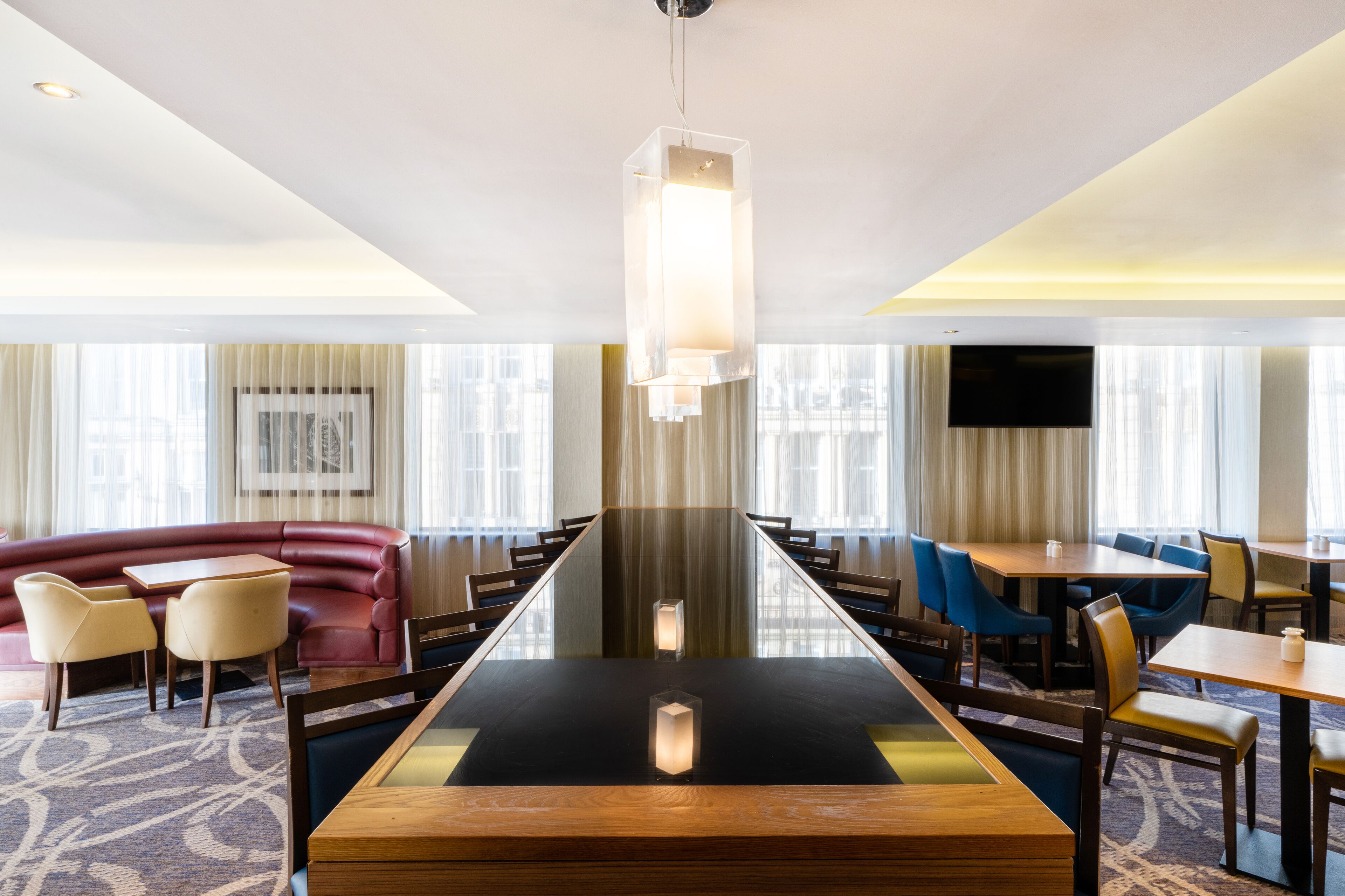 A modern hotel dining area with a long rectangular table, comfortable seating, and a large flat-screen TV mounted on the wall.