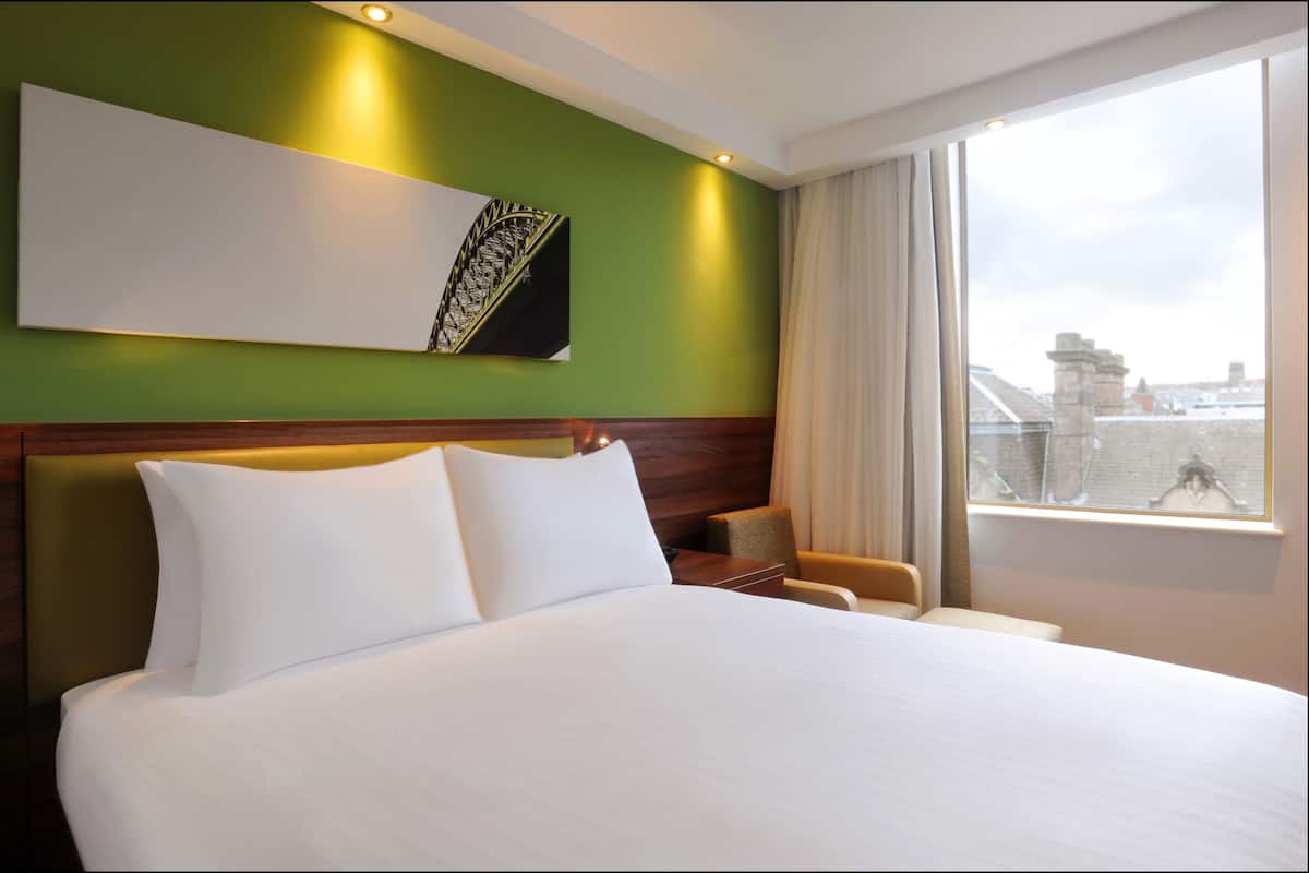 Ein Hotelzimmer mit einem großen Bett, einem Kopfteil, einem Wandbild, einem Fenster mit Vorhängen und Blick auf die Stadtlandschaft.