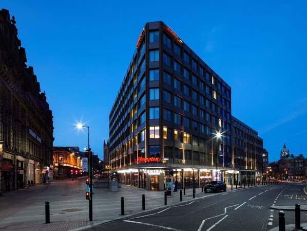 A modern hotel with a corner building, large windows, and a well-lit entrance. - Hampton By Hilton Newcastle (Newcastle-upon-Tyne)