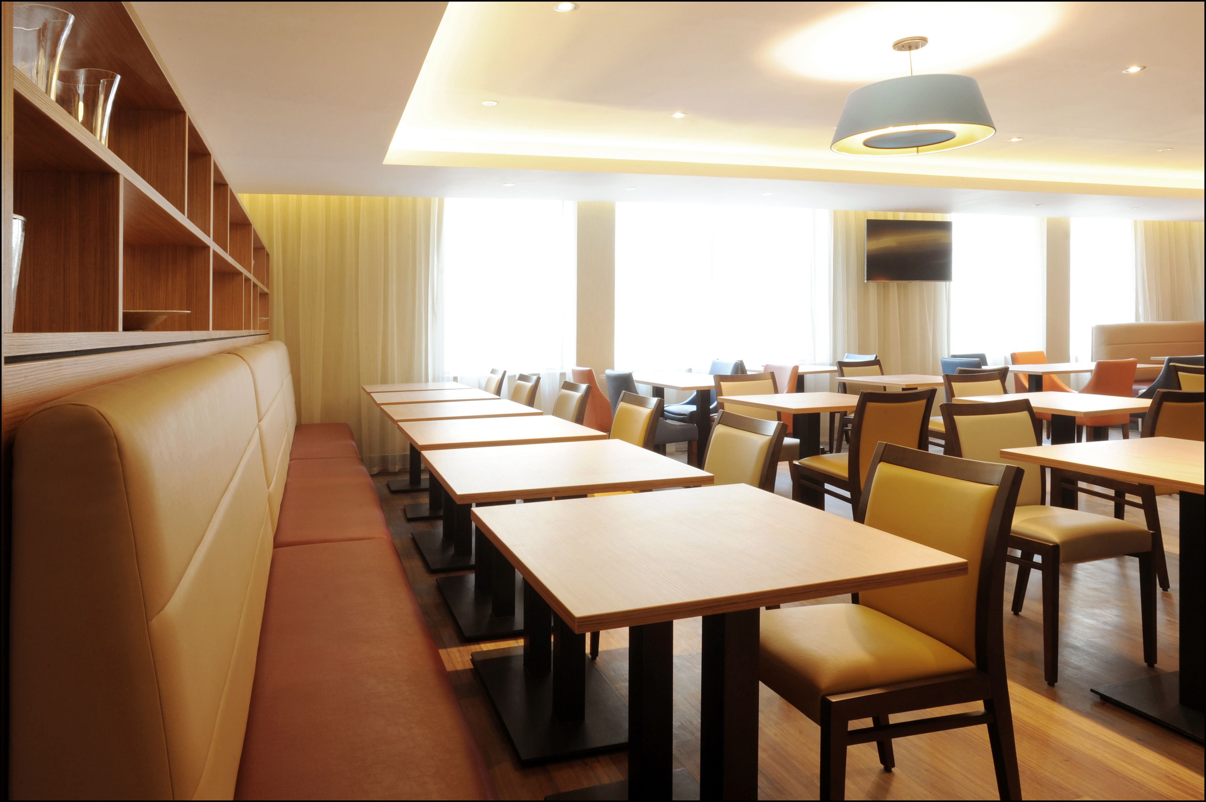 En restaurang med långa träbord och bekväma sittplatser, stora fönster och en modern taklampa.