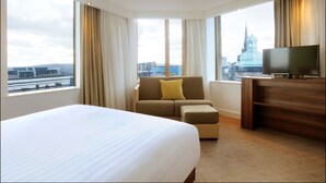 Ein Hotelzimmer mit einem großen Bett, einer Sitzecke, einem Fernseher und einem Schreibtisch. Durch große Fenster bietet sich ein Blick auf die Stadt.